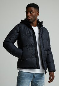 Chaqueta acolchada en negro, con capucha y cierre en la parte frontal. Patrón texturizado visible en la tela. Lleva puesta sobre una camisa blanca con jeans desgastados.