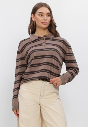 Frau trägt ein braun und dunkel gestreiftes Langarm-Poloshirt, in hellbeige Hose mit schwarzem Gürtel gesteckt, steht vor einfachem Hintergrund.