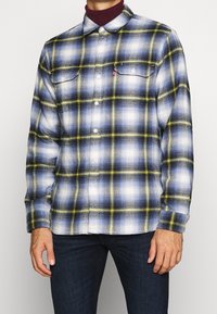 Camicia da uomo in flanella con un motivo a quadri blu, bianco e giallo, design a bottoni e due tasche sul petto. Tessuto morbido e maniche lunghe.