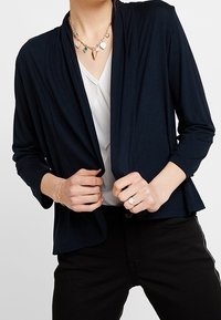 Marinblå cardigan med trekvartslång ärm, lättviktigt tyg, öppet frontdesign, lagrad över en vit topp, matchad med svarta byxor.