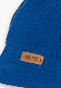 Blå bøttehatt laget av teksturert stoff, med en brun lærmerke som har "PURE PURE" preget. Enkel design med en nedadgående brem.