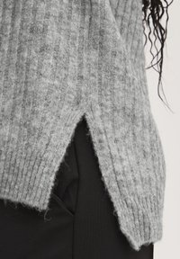 Grauer, gerippter Strickpullover mit seitlichen Schlitzen, aus einem weich strukturierten Material; trägt sich über einem schwarzen Kleidungsstück. Die Kanten sind leicht uneben.