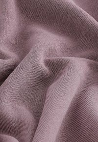 Měkká, texturovaná tkanina v tlumené fialové barvě, s jemnými pletenými detaily a jemným padajícím záhybem vytvářejícím hladké, plynulé ohyby.
