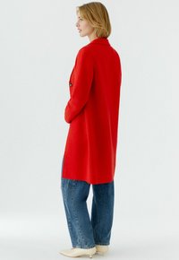 Long red coat with a collar, single button closure, textured fabric, and side pockets, paired with relaxed blue jeans and beige shoes.