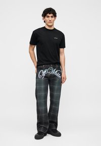Νέος άνδρας που φοράει μαύρο μπλουζάκι Off-White και σκούρο καρό παντελόνι με μεγάλο κείμενο "Off-White" στο μπροστινό μέρος.