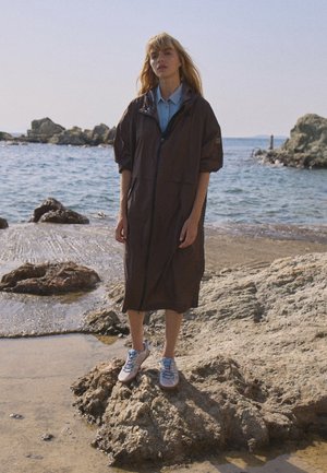 Femme debout sur une rive rocheuse portant un long manteau foncé, une chemise bleue et des baskets, avec la mer et des rochers en arrière-plan sous un ciel clair.