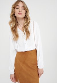 Witte button-up blouse van gestructureerde stof, met lange mouwen en gedetailleerde naden, gecombineerd met een bruine suedewraprok met een zijsplit.