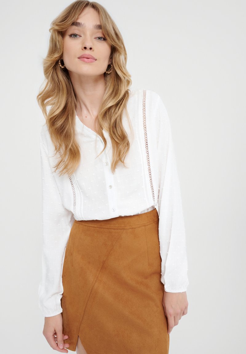 Witte button-up blouse van gestructureerde stof, met lange mouwen en gedetailleerde naden, gecombineerd met een bruine suedewraprok met een zijsplit.