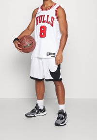 Canotta da basket bianca con grafica rossa "BULLS" e numero 8, abbinata a pantaloni corti bianchi con bordi neri. Scarpe da basket Nike nere e grigie.