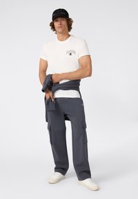Hombre con gorra negra, camiseta blanca de Champion, pantalones cargo grises, zapatillas blancas, sosteniendo un sudadera gris a juego alrededor de la cintura.