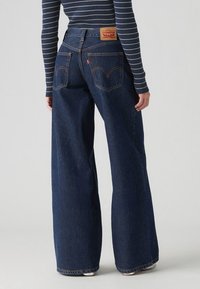 Jeans en denim à jambe large de couleur bleu foncé, avec une taille mi-haute, deux poches arrière et un patch en cuir de marque sur la ceinture.