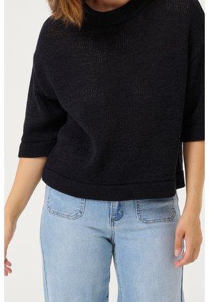 Femme portant un pull noir en maille à manches courtes et un jean denim bleu clair, montrée du cou aux hanches sur un fond uni.