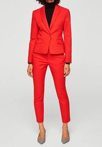 Červený oblek na míru se zapínáním blazru na jeden knoflík a úzkými kalhotami. Dokon�čeno šedými podpatky.