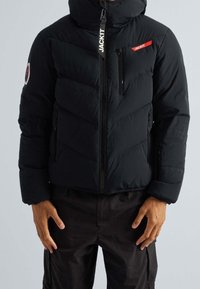 Doudoune noire matelassée avec fermeture éclair à l'avant, capuche et couture en chevrons. Comprend une poche latérale et un patch logo sur la manche. Texture douce.