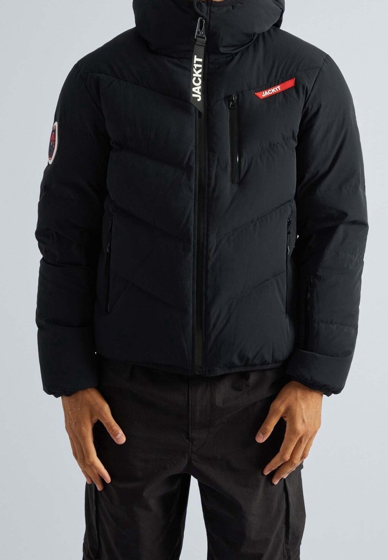 Doudoune noire matelassée avec fermeture éclair à l'avant, capuche et couture en chevrons. Comprend une poche latérale et un patch logo sur la manche. Texture douce.