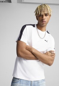 Camiseta blanca de manga corta con acentos negros en los hombros y un pequeño logo negro. Material de algodón con un corte relajado, combinada con jeans de lavado claro.