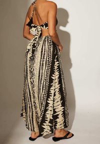 Femme portant un top dos nu à col halter et une longue jupe à motifs en noir et beige, debout les mains sur les hanches, portant des sandales noires.