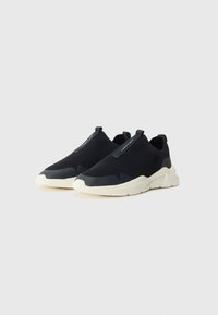 Crne pletene slip-on tenisice s teksturiranim gornjim dijelom, bijelim gumenim potplatom i trakom na peti za lako obuvanje/izvlačenje.