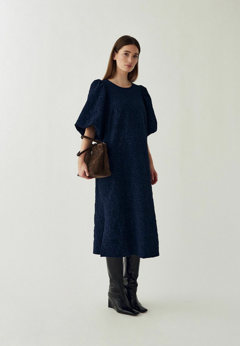 Femme debout portant une robe midi texturée bleu marine à manches larges, des bottes noires hauteur genou et tenant un sac bandoulière en daim marron.