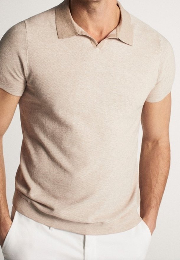 Homme portant un polo beige à manches courtes rentré dans un pantalon blanc, les mains dans les poches, sur un fond uni.