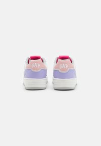 Zapatillas blancas con acentos en púrpura y rosa en el talón y los laterales. Cuenta con una tela texturizada y una lengüeta en la parte posterior. Logo "GAP" en el talón.