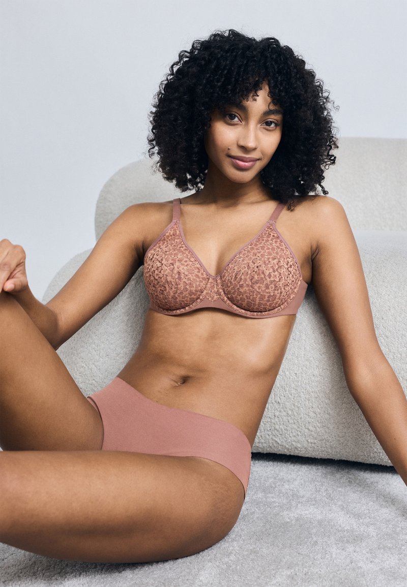 Γυναίκα με κατσαρά μαλλιά καθισμένη σε δάπεδο με χαλί, φορώντας ταιριαστό δαντελένιο σουτιέν σε μωβ αποχρώσεις και αόρατα εσώρουχα, μπροστά σε ουδέτερο φόντο.