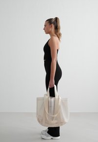 Kassi läpinäkyvä beige, jossa on kermaiset hihnat, suorakaiteen muotoinen ja avoin yläosa, jota pitelee henkilö mustissa urheiluvaatteissa ja valkoisissa lenkkareissa.