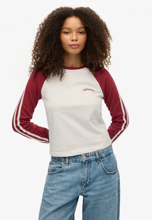 Camiseta de manga larga blanca y marrón con el logo "Superdry", mangas raglán y dos rayas blancas en las mangas, combinada con unos jeans de mezclilla claros.