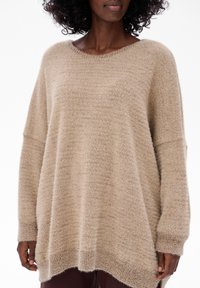 Beżowy sweter dziany o luźnym kroju, szerokim dekolcie w łódkę i długich rękawach. Miękka, puszysta tekstura z prążkowanymi mankietami i dłuższym tylnym dołem.