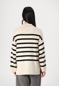 Suéter a rayas beige con líneas horizontales negras, con un corte holgado, mangas largas y un cuello de chal. Textura suave, diseño de longitud media.