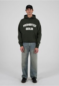 Tânăr purtând o șapcă neagră, hanorac verde închis cu textul „PROHIBITED BERLIN”, blugi albaștri și pantofi negri, stând în fața unui fundal simplu.