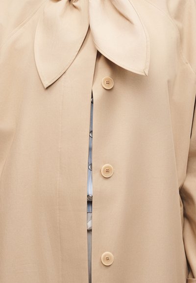 Μπεζ παλτό με μεγάλο γιακά σε σχήμα φιόγκου και τρία ορατά στρογγυλά κουμπιά, που αποκαλύπτει εν μέρει μια μπλε και λευκή ένδυση από κάτω.