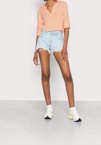 Blusa arancione a righe con scollatura a V, abbinata a pantaloni corti di jeans chiari con orli sfrangiati. Indossata con sneakers bianche e giallo neon.