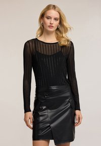 Schwarzes Langarmoberteil aus transparenter Stoff mit vertikalen Streifen, kombiniert mit einem schwarzen Leder-Minikleid, ausgestattet mit Druckknöpfen und vorderen Taschen.