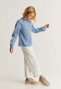 Cămașă butonată de culoare bleu deschis, cu detalii brodate pe umeri, asortată cu pantaloni albi scurți și adidași rose gold. Textura materialului este netedă.