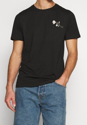 Mann trägt ein schwarzes, kurzärmeliges T-Shirt mit kleinem weißen Blumenmotiv auf der Brust und blaue Jeans, Hand in der Tasche.