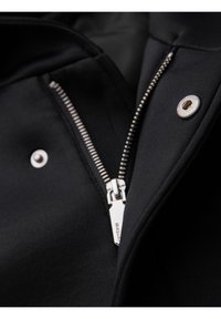 Gros plan sur une veste noire partiellement zippée avec une fermeture éclair argentée et des boutons-pression ronds en argent sur un tissu lisse.