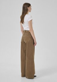 Pantalones de pierna ancha marrones hechos de un tejido texturizado, con una cintura alta, bolsillos traseros y detalles de costura visibles. Combinados con una camisa blanca.