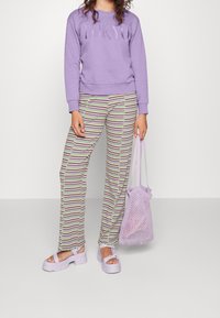 Giovane donna che indossa un felpa lavanda con la scritta "TOKYO", pantaloni a strisce, sandali con suola piatta e tiene in mano una borsa a rete lavanda.