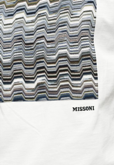 Λευκό ύφασμα με πολύχρωμο ζιγκ-ζαγκ σχέδιο κεντημένο, με τη λέξη «MISSONI» κεντημένη με μαύρα γράμματα κάτω από το σχέδιο.