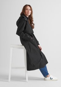 Black waterproof trench coat with a hood, cinched waist, and snap closures. Worn over blue jeans and white sneakers.