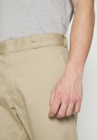Male arm and hand resting next to beige pants pocket, with a light grey shirt tucked into the waistband.