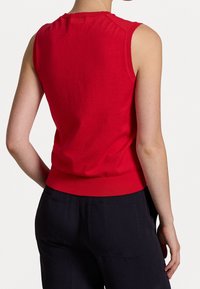 Person, der ein ärmelloses rotes Oberteil und schwarze Hose trägt, von hinten vor einem weißen Hintergrund gezeigt.