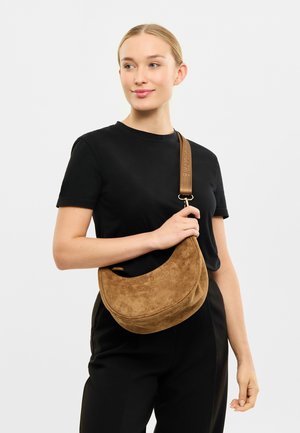 Hnědá semišová taška přes rameno se zakřiveným tvarem a širokým popruhem s embosovaným písmem. Taška má jemnou texturu a minimalistický design.