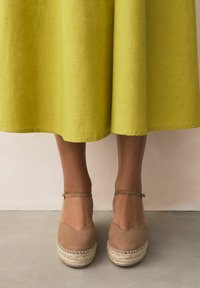 Espadrillas in camoscio marrone con design a punta a V, suole in juta e cinturini alla caviglia, abbinate a una gonna gialla fino al ginocchio.