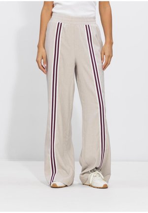 Beige broek met wijde pijpen, elastische tailleband en dubbele verticale strepen in bordeauxrood en wit, gedragen met witte sneakers en een wit shirt.
