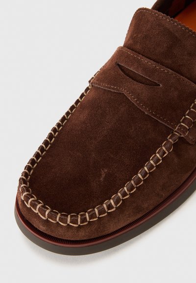 Mocassins en daim marron avec un design à enfiler, des coutures contrastantes et une découpe décorative près du cou-de-pied. Semelle en caoutchouc plate.