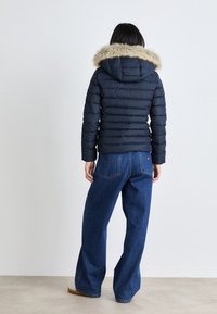 Navy puffer jacket with a faux fur hood, featuring a quilted design. Paired with wide-leg blue jeans. The back view shows a sleek silhouette.