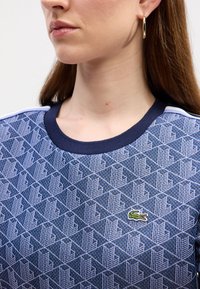 Bordo zila džemperis ar ģeometrisku rakstu gaišāk zila krāsā, ribināta apaļa kakla izgriezums un neliels zaļš krokodila logotips uz krūtīm.