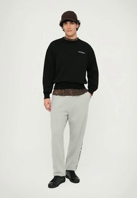 Black sweatshirt with logo, layered over a brown patterned shirt, paired with light grey sweatpants and black sneakers. Model stands against a plain background.